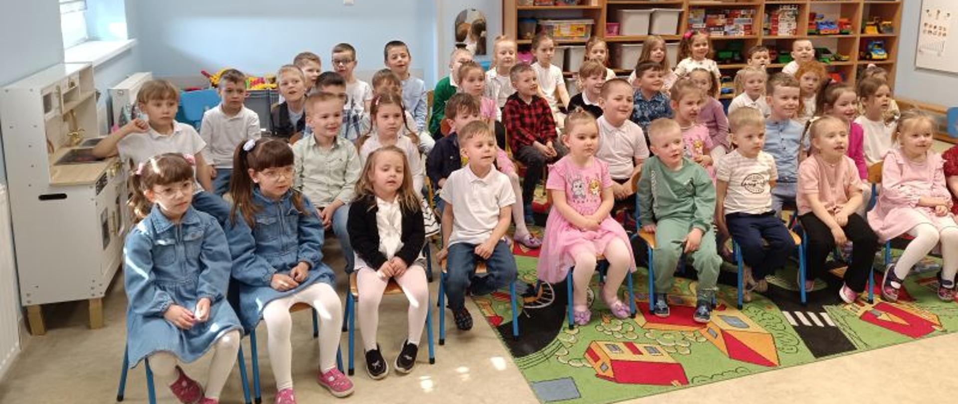 Grupa dzieci wieku przedszkolnym siedzi na małych krzesłach. Na podłodze leży kolorowy dywan, za dziećmi widać półki z zabawkami, książkami. Ściany sali mają kolor niebieski, po lewej stronie widać okno z roletami. Na suficie świetlówki.