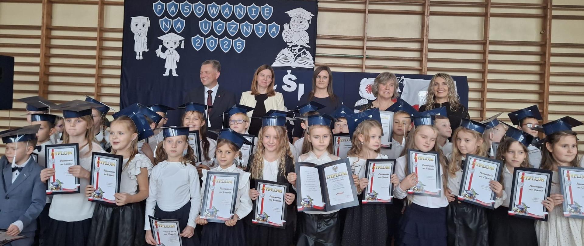 Pierwszoklasiści z dyplomami w odświętnych strojach. Za uczniami stoją Wójt Gminy Mielec, Dyrekcja Zespołu Szkół w Chorzelowie i wychowawcy klas pierwszych.