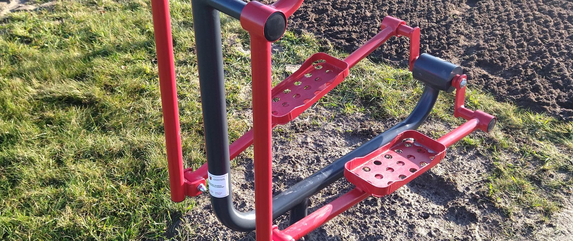 Obraz przedstawia plenerowy orbitrek treningowy zainstalowany na trawiastym terenie. Maszyna ma czerwone uchwyty oraz pedały, a jej centralna konstrukcja jest czarna. W tle widoczny jest ogrodzony obszar z zaoraną ziemią, zaparkowany samochód oraz kilka budynków.