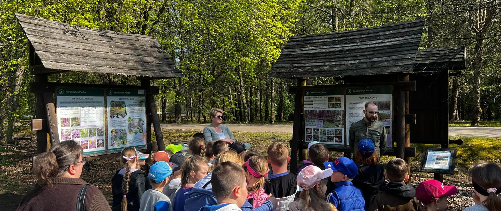 Dzieci wraz z leśnikiem znajdują się w lesie na ścieżce edukacyjnej. Uczniowie skierowani są w stronę Pana leśnika. Za nim stoją tablice informacyjne.