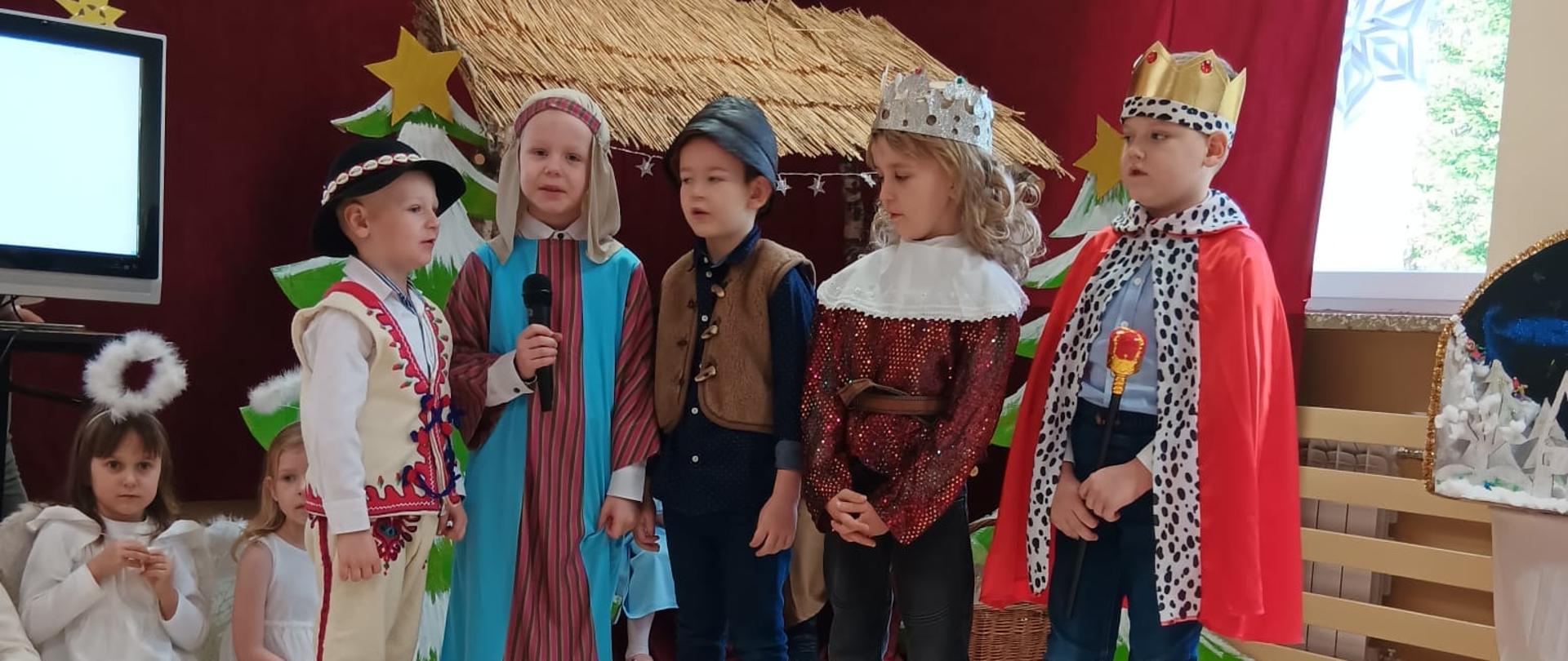 Przedszkolaki odgrywają jasełka w przebraniach świątecznych, w tle szopka