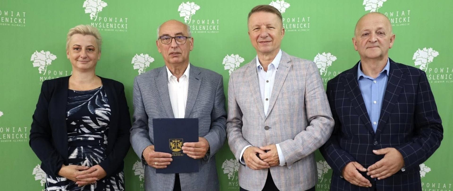 Na zdjęciu widzimy cztery osoby stojące na tle ścianki promocyjnej z logo i napisem Powiat Myślenicki Ziemia o Dobrym Klimacie. Tło w kolorze zielonym zawiera również biały kontur mapy powiatu. Osoby ustawione są w szeregu i patrzą w stronę aparatu. Wszyscy są elegancko ubrani, dwie kobiety i dwóch mężczyzn mają na sobie marynarki. Mężczyzna drugi od lewej trzyma w dłoniach granatową teczkę z herbem powiatu myślenickiego. Zdjęcie zostało wykonane przy okazji podpisania umowy. Atmosfera jest formalna, ale przyjazna, osoby się uśmiechają. Zdjęcie dokumentuje współpracę lokalnych przedstawicieli samorządu.