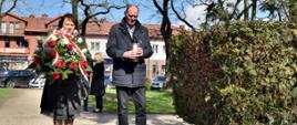Na fotografii starosta Elżbieta Sadowska oraz wicestarosta Ryszard Domański stojący przed pomnikiem. Starosta trzyma w dłoniach wiązankę z biało - czerwonych kwiatów i patriotycznymi szarfami w kolorach flagi państwowej. Obok wicestarosta Domański w dłoniach trzyma znicz, który za chwile zostanie złożony ku pamięci ofiar katastrofy smoleńskiej.