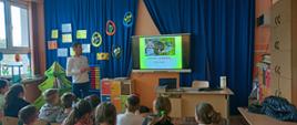 Mistrz w biegach pan Piotr Prusak wyświetla na ekranie dotykowym prezentację o swoich dokonaniach, przed nim siedzą uczniowie, z tyłu klasy widoczne szafki szkolne