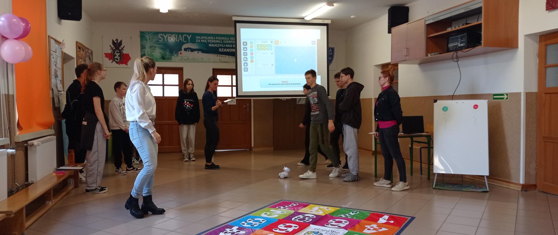 Grupa uczniów wraz z trzema nauczycielkami stoją na korytarzu. Między nimi stoi robot Photon, obok niego leży kolorowa mata z liczbami.