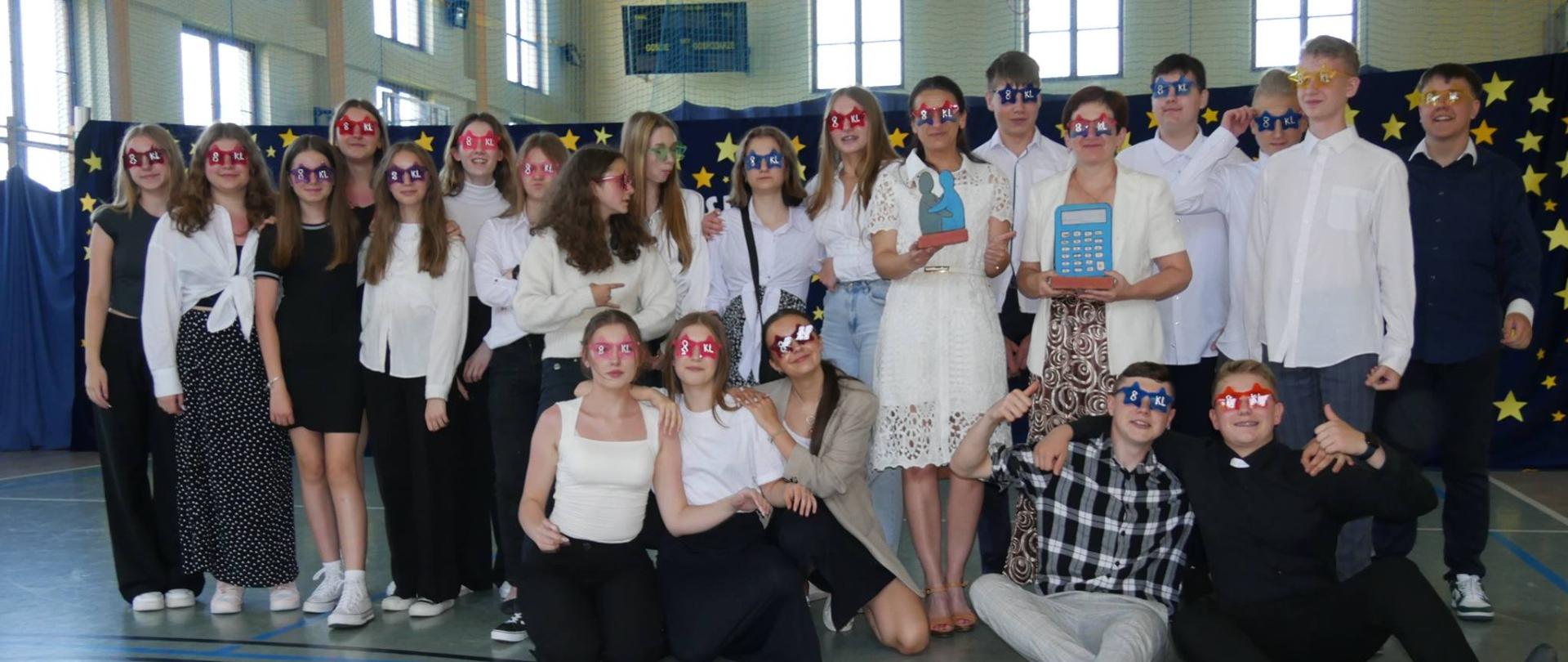 Absolwenci klas VIII wraz z wychowawcom i nauczycielem współorganizującym proces kształcenia na tle pięknej dekoracji w kolorowych okularach z napisem klasa VIII
