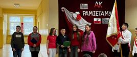 Trzy uczennice i jeden uczeń śpiewają pieśń, po prawej stronie stoi poczet sztandarowy szkoły ze sztandarem, za nimi bordowa zasłona, na której przypięto biało czerwoną flagę, napis "AK pamiętamy" oraz symbol Polski walczącej, na pierwszym planie widać uczniów oglądających akademię