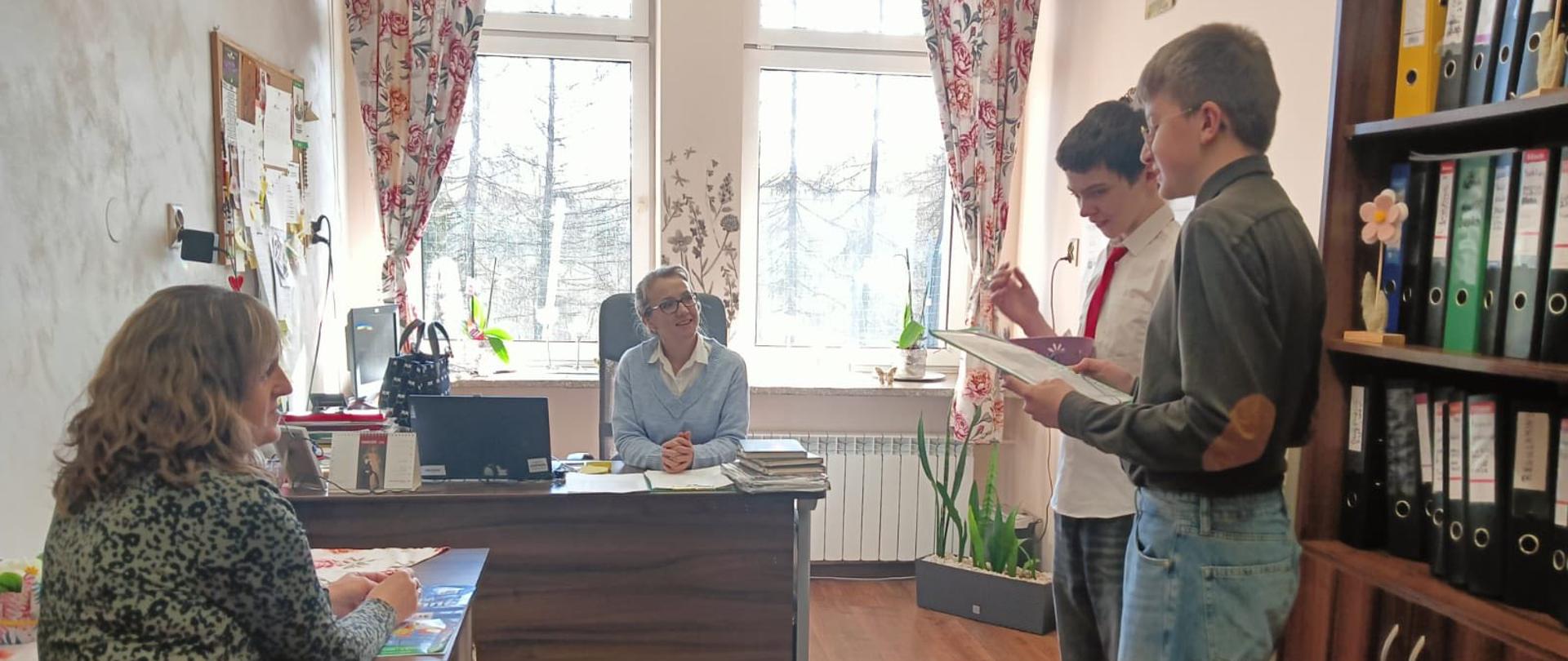 Dwóch chłopców przedstawicieli Samorządu Uczniowskiego częstuje składa życzenia pani dyrektor w gabinecie dyrektorskim i siedzącej tam pani pedagog