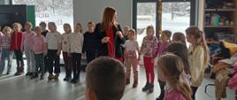 Dzieci biorą udział w regionalnej zabawie, o której pisze pisarka w swoich książkach, stoją w kole i przyglądają się, jak pisarka nawija na sznurek wirującą zabawkę