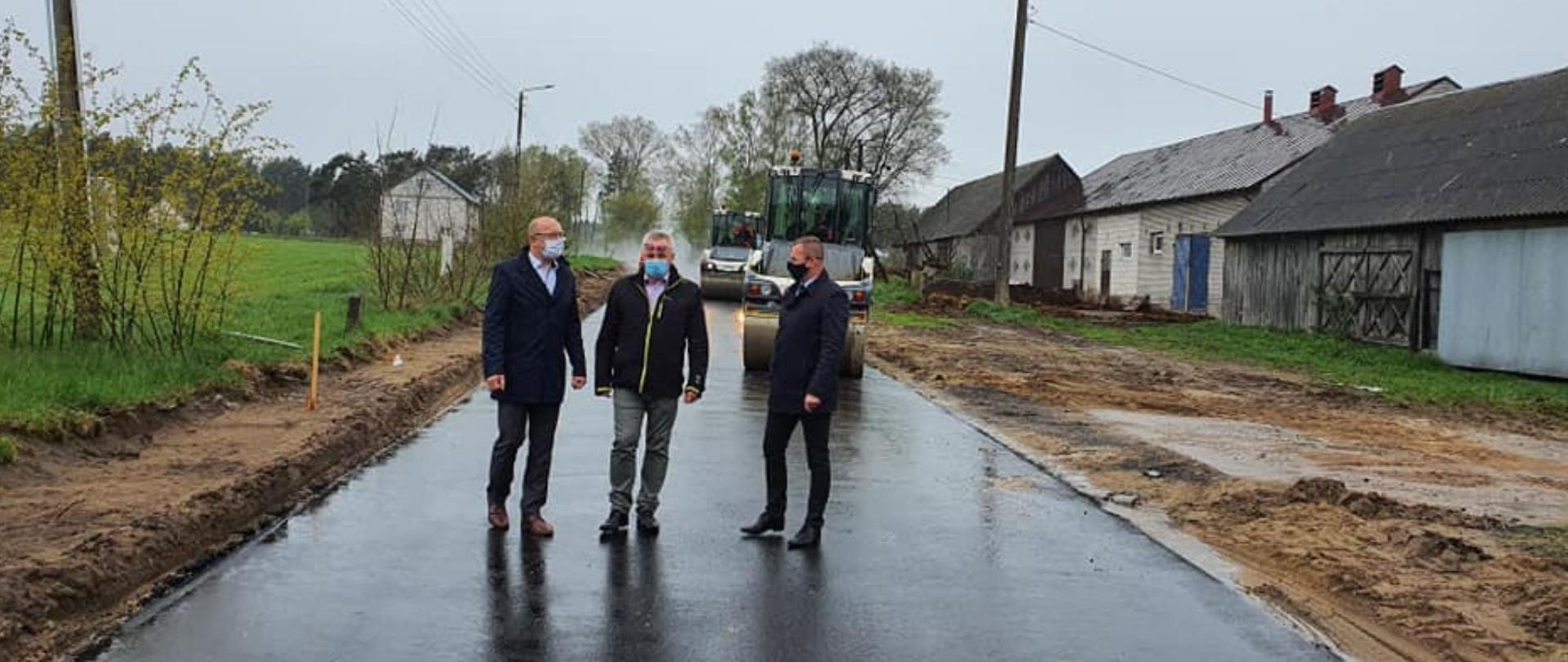 na zdjęciu znajdują się trzy osoby, które stoją na drodze, w tle dwa walce maszynowe