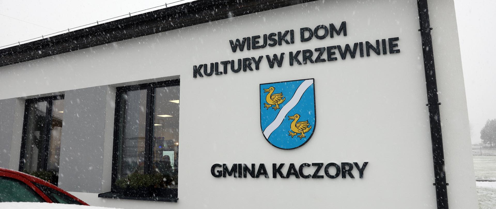 Na zdjęciu widać wiejski dom kultury w Krzewinie wraz z napisem i herbem gminy