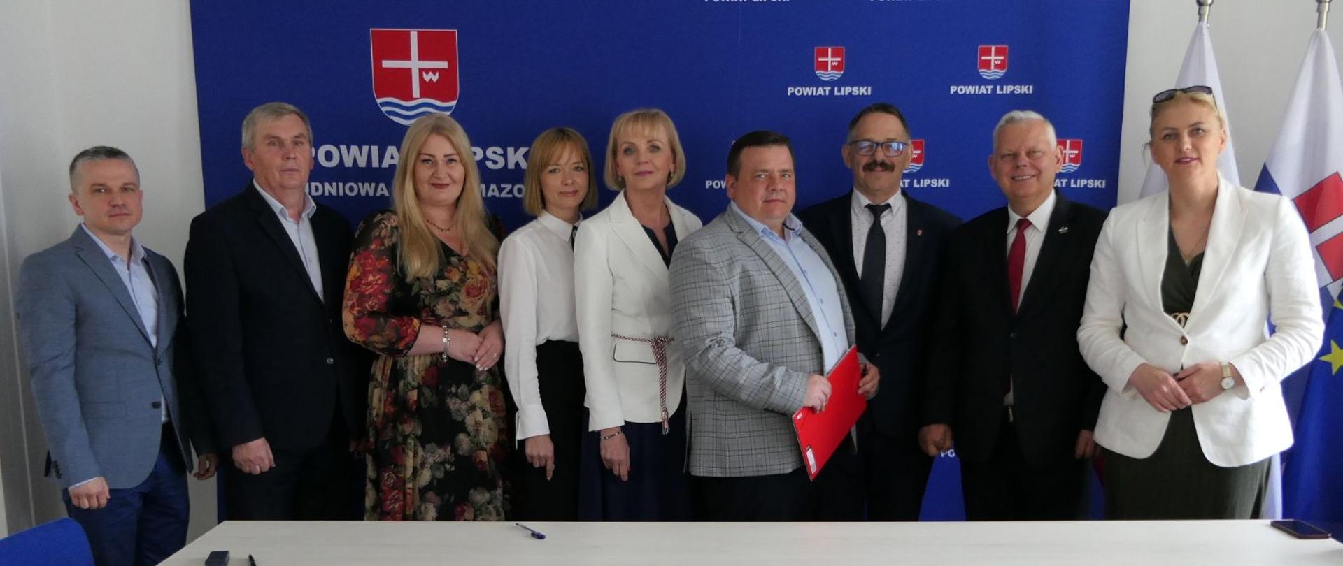 Uczestnicy wydarzenia przed podpisaniem umowy. W tle ścianka granatowa ścianka z herbem powiatu.