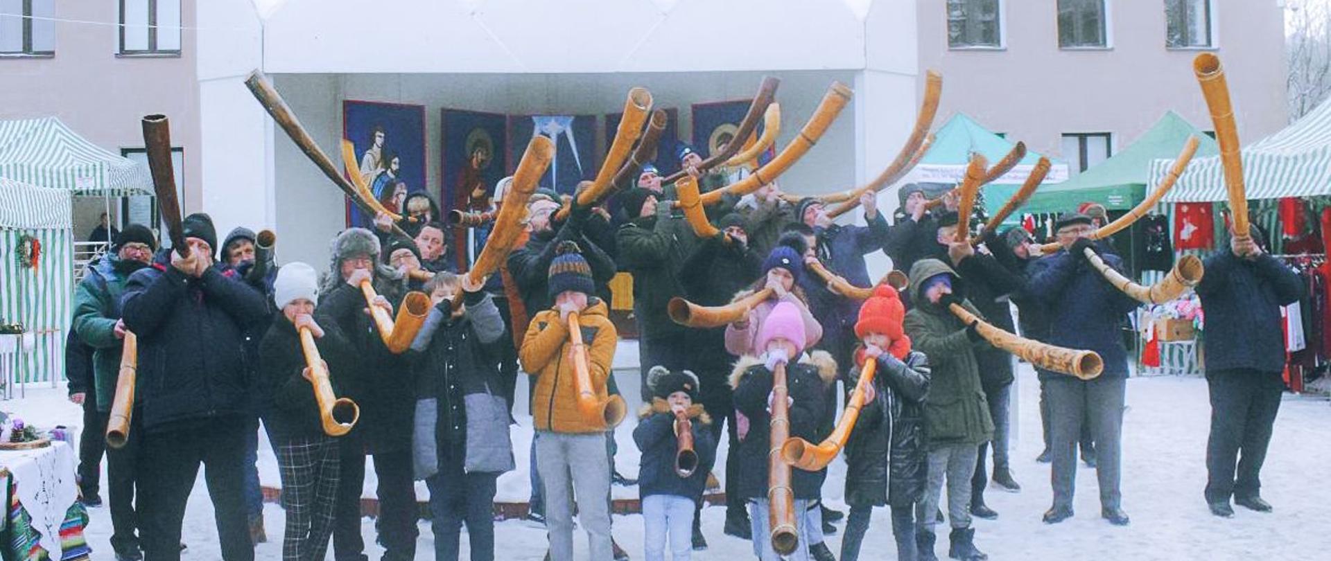 Zdjęcie grupowe. Na tle gmachu Sokołowskiego Ośrodka Kultury i stojącej przed nim szopki bożonarodzeniowej uczestnicy prezentacji gry na ligawkach. Dmuchają w skierowane ku górze długie zagięte drewniane trąby (ligawki).