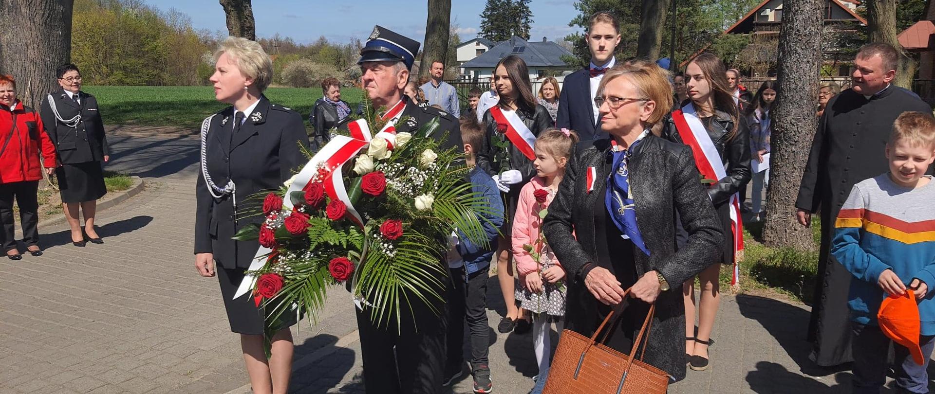 Uczestnicy obchodów przed pomnikiem w pozycji zasadniczej do hymnu. na pierwszym planie trzy osoby, w środku strażak z wiązanką kwiatów, oraz dwie panie. Za nimi dzieci z kwiatami w ręku, następnie młodzież z szarfami w kolorach biało-czerwonych. W głębi pozostali uczestnicy uroczystości.