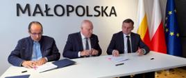 Trzech mężczyzn siedzi przy stole podczas podpisania dokumentów w siedzibie Małopolski, w tle widoczne są flagi Polski i Unii Europejskiej.