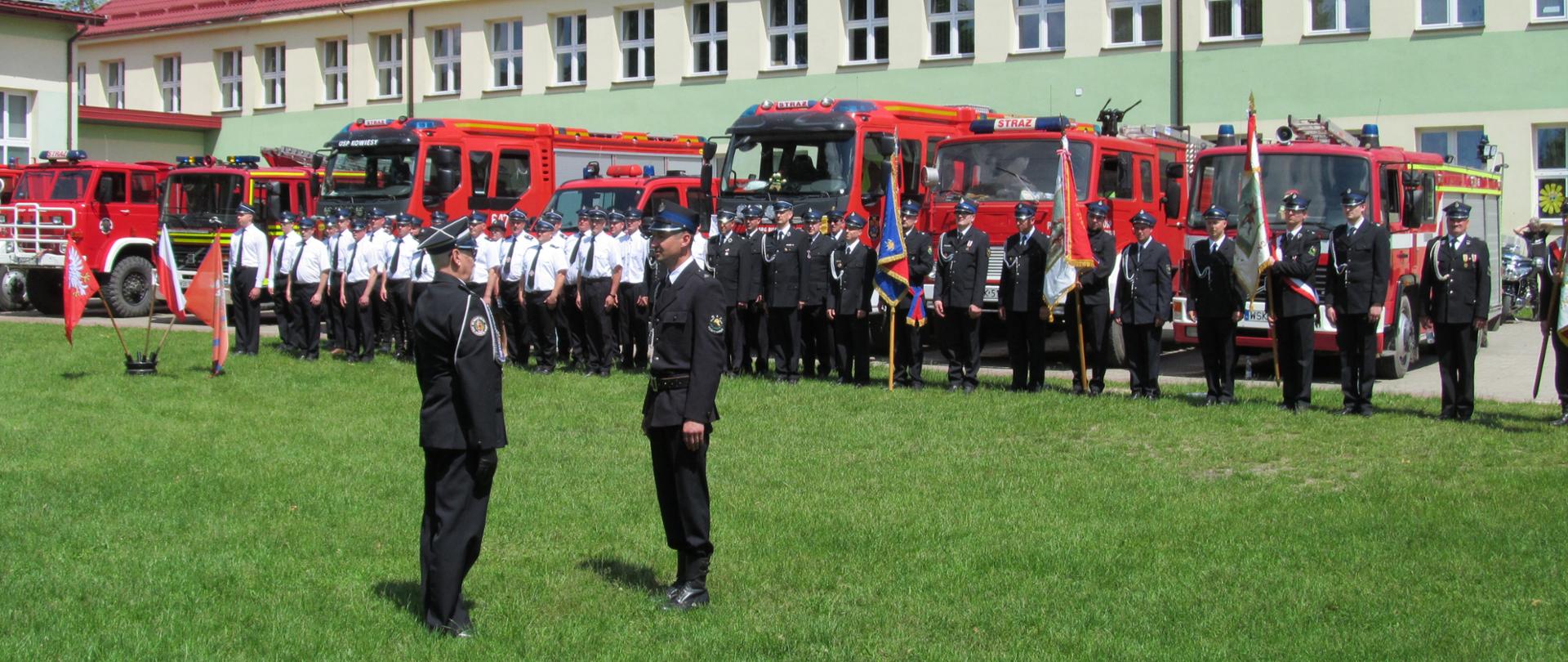 Uroczysta zbiórka. Na środku dużego trawnika strażak zdaje meldunek dowódcy. W tle ustawieni w szeregu strażacy zaproszonych jednostek OSP, stojące obok siebie poczty sztandarowe, wozy strażackie oraz budynek.