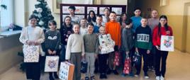 Grupa uczniów nagrodzonych w konkursie "Świąteczny Stroik Bożonarodzeniowy" stoi na korytarzu szkolnym, obok choinki, uczniowie trzymają w ręku ozdobne papierowe torby z nagrodami, na ścianie w gablocie wisi sztandar szkoły przedstawiający patronkę Świętą Kingę