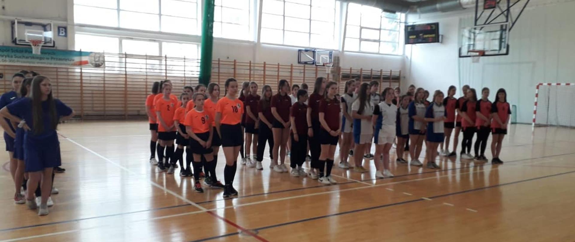Na Hali Sportowej w Przasnyszu rozegrano finały Powiatowych Igrzysk Młodzieży Szkolnej w Koszykówce dziewcząt w kategorii Szkół Podstawowych. Zdjęcie przedstawia dziewczęta rozstawiona jako drużyny.