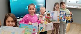 Uczniowie klasy 1 trzymają w ręku wykonane przez siebie plakaty o Smerfach, za nimi monitor interaktywny