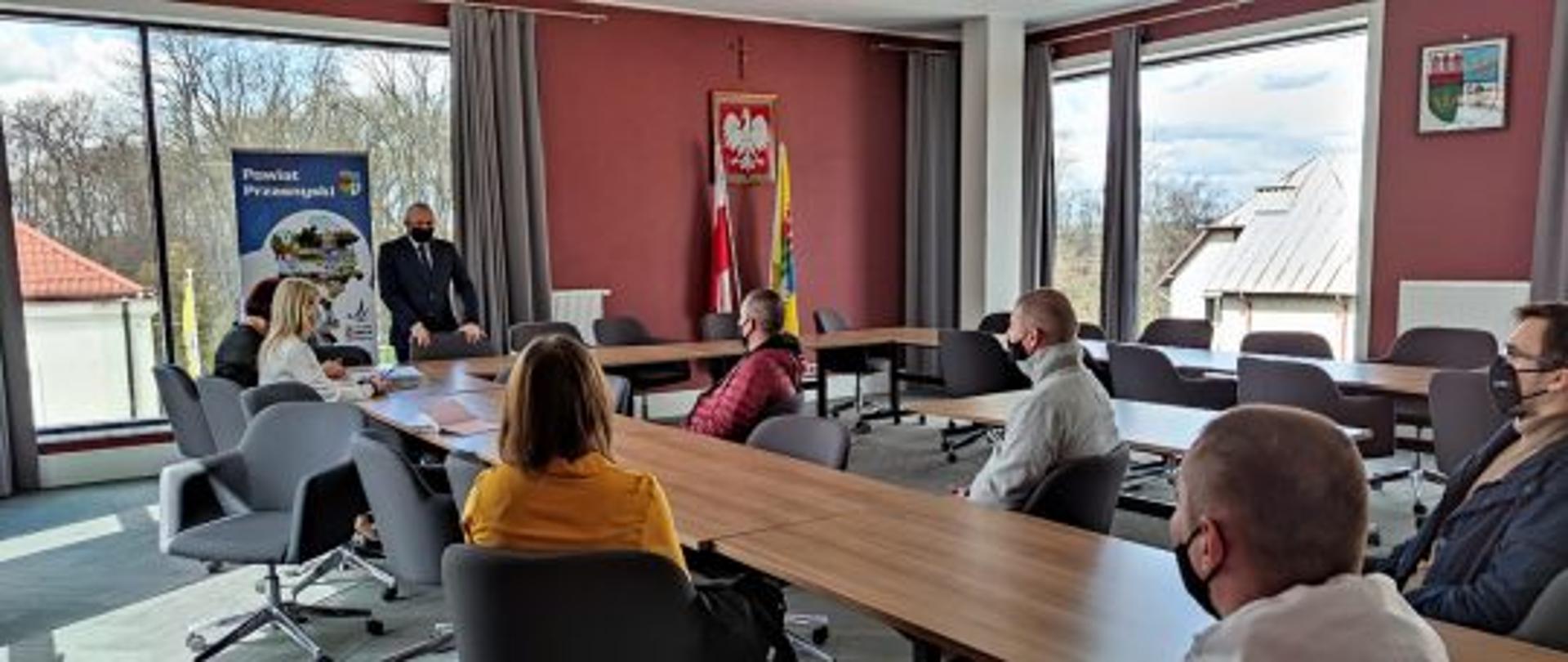 Zdjęcie przedstawia Starostę Przasnyskiego Krzysztofa Bieńkowskiego, Wicedyrektor Powiatowego Urzędu Pracy Annę Obrębską oraz Panią Barbarę Garlińską wraz z nowymi przedsiębiorcami powiatu przasnyskiego na chwilę przed podpisaniem umów na dofinansowanie rozpoczęcia ich działalności gospodarczej. Zdjęcie wykonano na sali konferencyjnej Centrum Aktywizacji Przedsiębiorczości w Starostwie Powiatowym w Przasnyszu, w tle szare kotary, ściany w kolorze szlachetnego mahoniu oraz oprawione godło Rzeczypospolitej Polskiej – biały orzeł w złotej koronie na czerwonym tle.