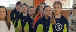 Drużyna uczennic w strojach granatowo-zielonych stoi w szeregu jedna za drugą obok drużyny przeciwnej