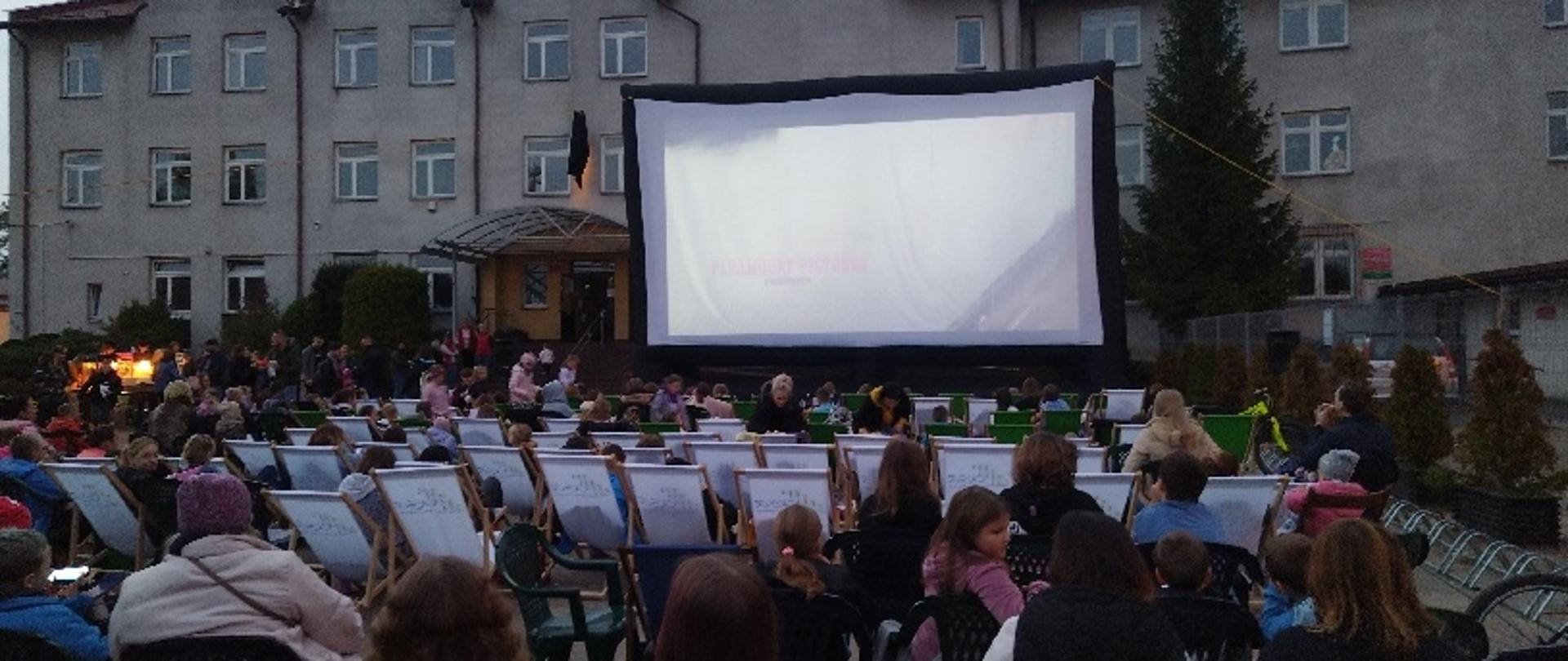 Ludzie siedzący na leżakach, oglądają film na dużym ekranie na placu przed dużym budynkiem