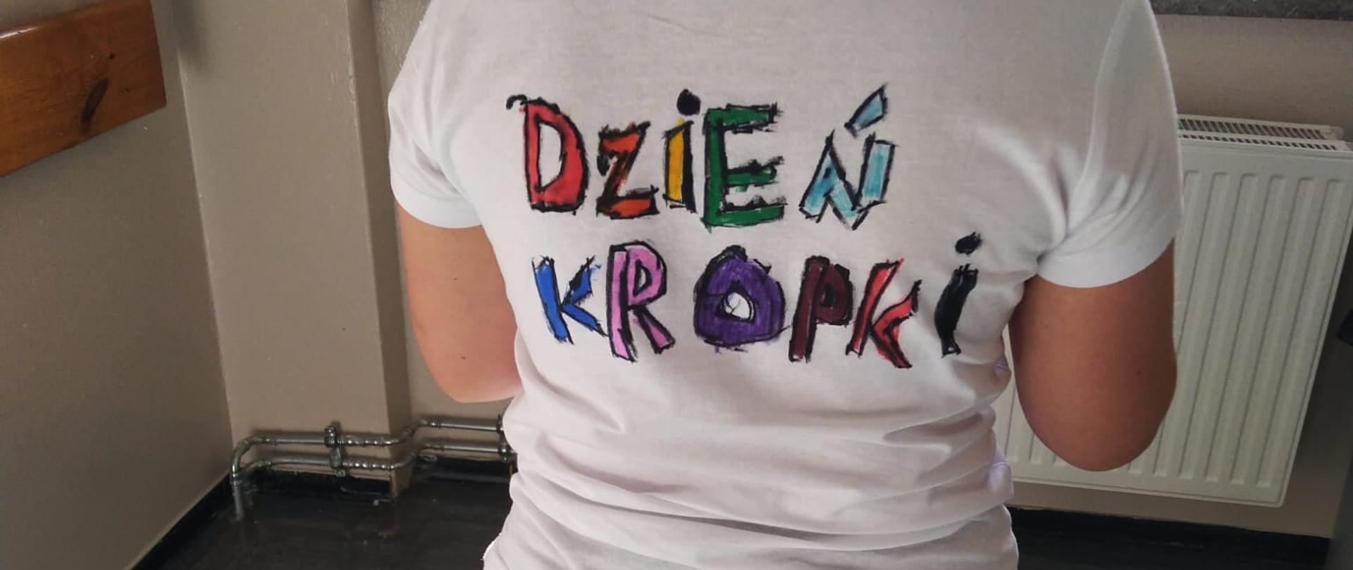 Na środku dziewczynka stojąca tyłem do zdjęcia. Ma ubraną białą koszulkę z kolorowym napisem dzień kropki. W tle widać okno.