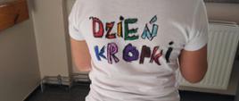 Na środku dziewczynka stojąca tyłem do zdjęcia. Ma ubraną białą koszulkę z kolorowym napisem dzień kropki. W tle widać okno.