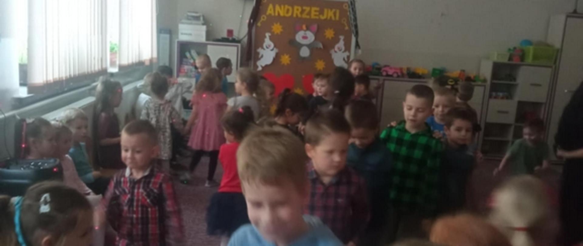 W sali przedszkolnej grupa dzieci tańczy do muzyki. W tle widoczny napis Andrzejki.