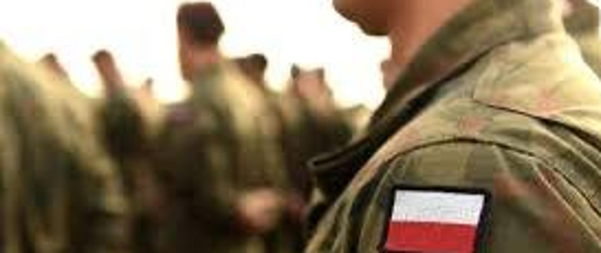 Żołnierz w mundurze z naszywką polskiej flagi na ramieniu, na tle innych żołnierzy