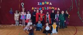 Uczniowie w przebraniach bajkowych stoją na sali gimnastycznej na tle przystrojonej bordowym płótnem ściany z kolorowym napisem "Zabawa andrzejkowa"