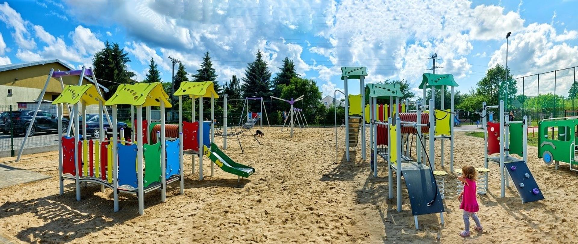 Plac zabaw wysypany żółtym piaskiem, na nim różnego rodzaju zabawki, domki, huśtawki i karuzele. Do jednego domku podchodzi dziewczynka w różowej sukience.
