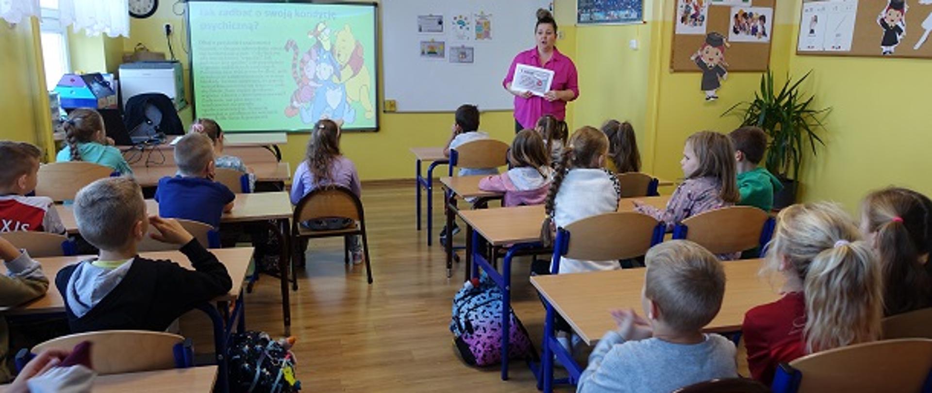 Dzieci klas młodszych siedzą w sali lekcyjnej. Nauczycielka prowadzi pogadankę.