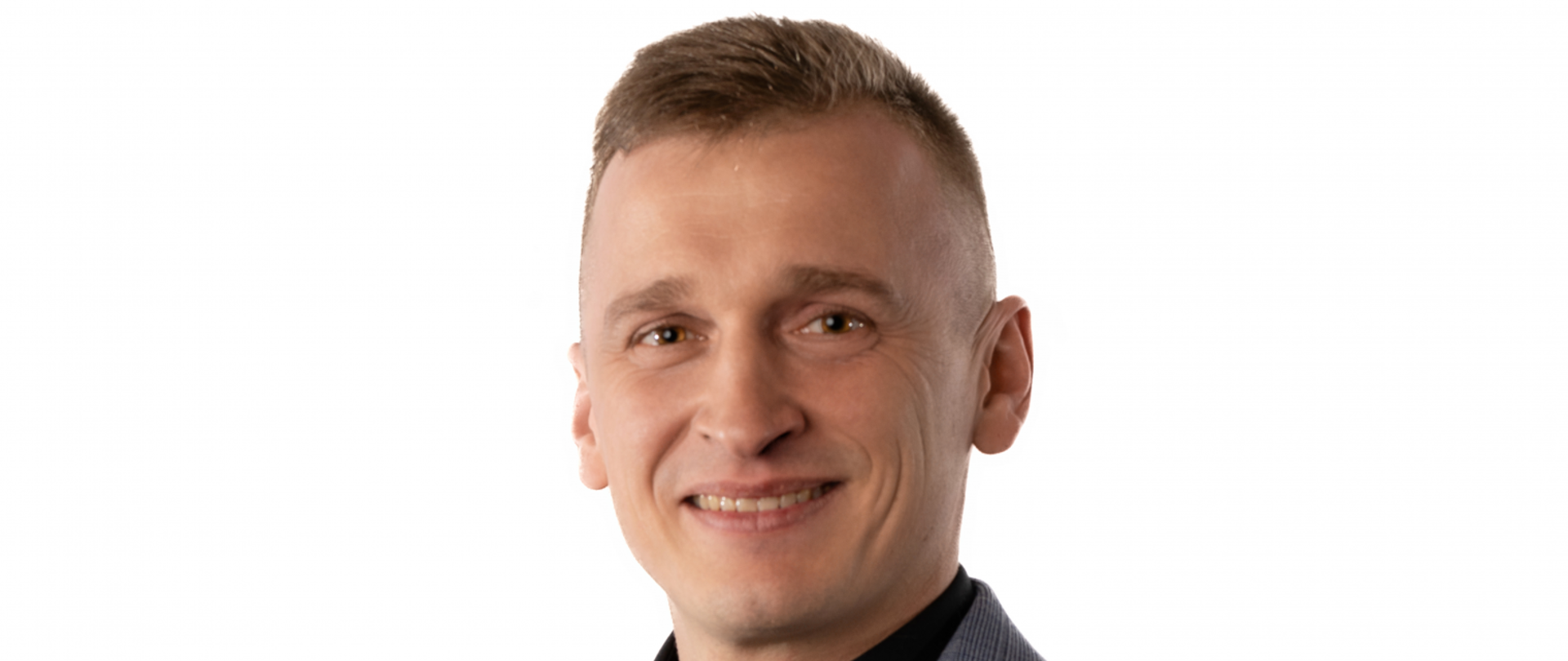 Szatyn, brązowe śmiejące się oczy, uśmiechnięta twarz. Ubrany w szary garnitur i czarna koszulę