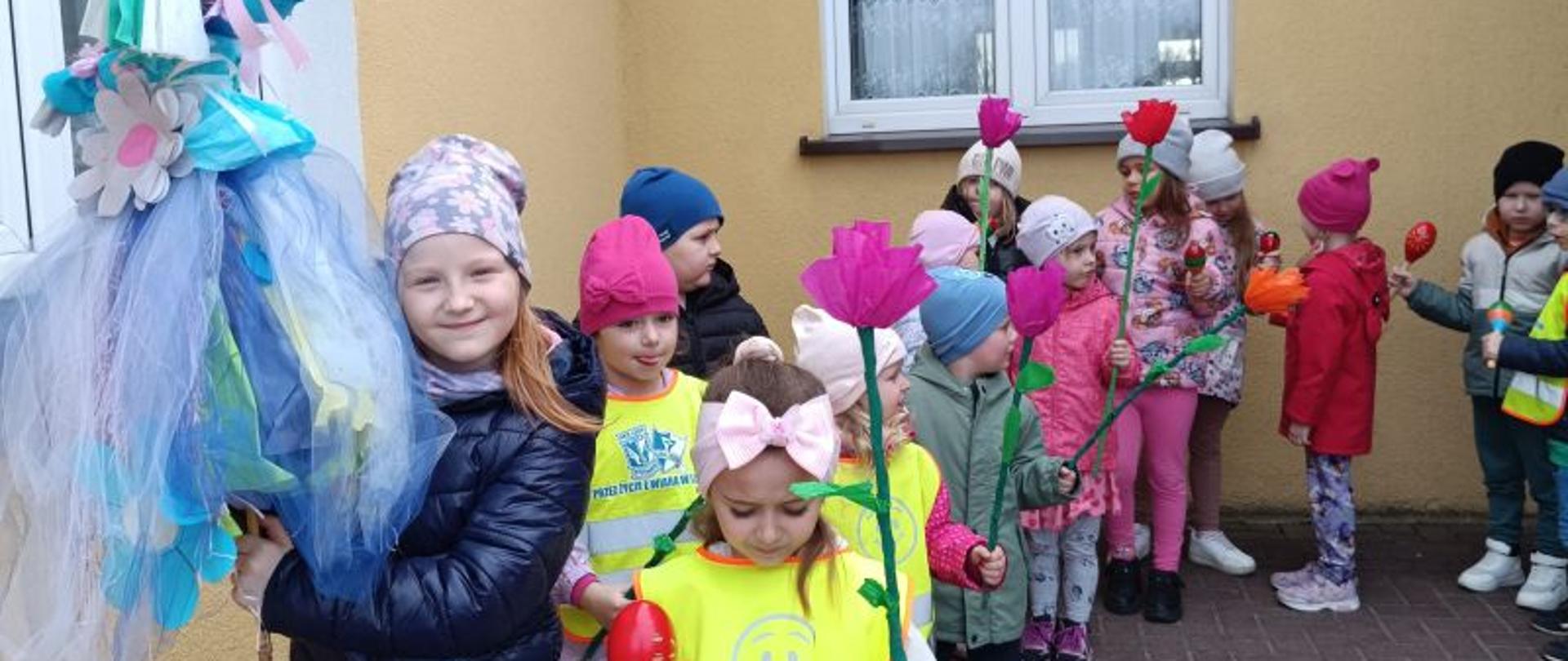 Grupa dzieci w wieku przedszkolnym w kurtkach i czapkach stoi przed budynkiem na chodniku, dziewczynka z przodu trzyma kukłę zrobioną z bibuły i kolorowej tkaniny. Niektóre dzieci mają założone kamizelki odblaskowe a w rękach trzymają kwiaty z bibuły. Po lewej stronie zdjęcia okno, w tle drugie okno budynku. 
