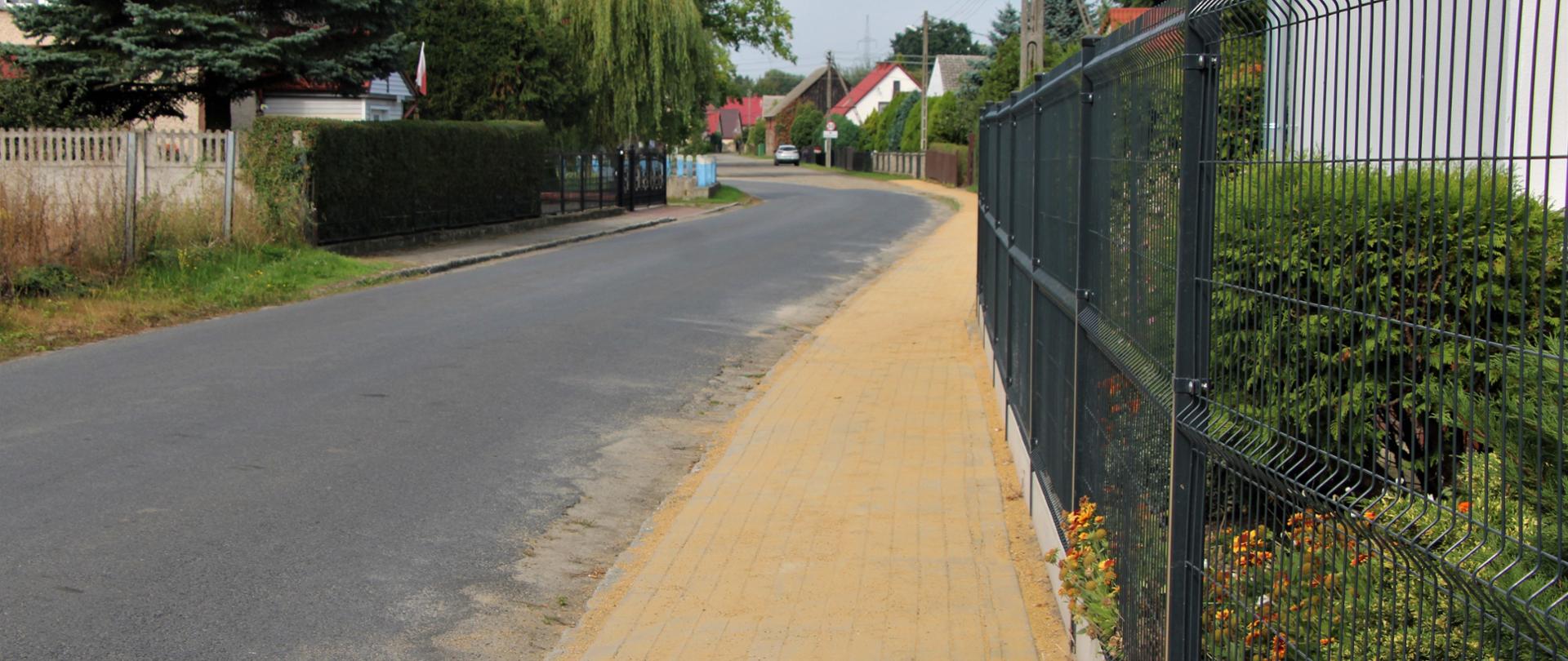 Droga asfaltowa i ścieżka z betonowej kostki, ogrodzenia oraz zabudowania we wsi. 