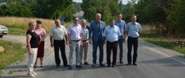 Na zdjęciu widać grupę ludzi stojących w rzędzie na jezdni. W tle widzimy drogę oraz po bokach rozciągają się pola