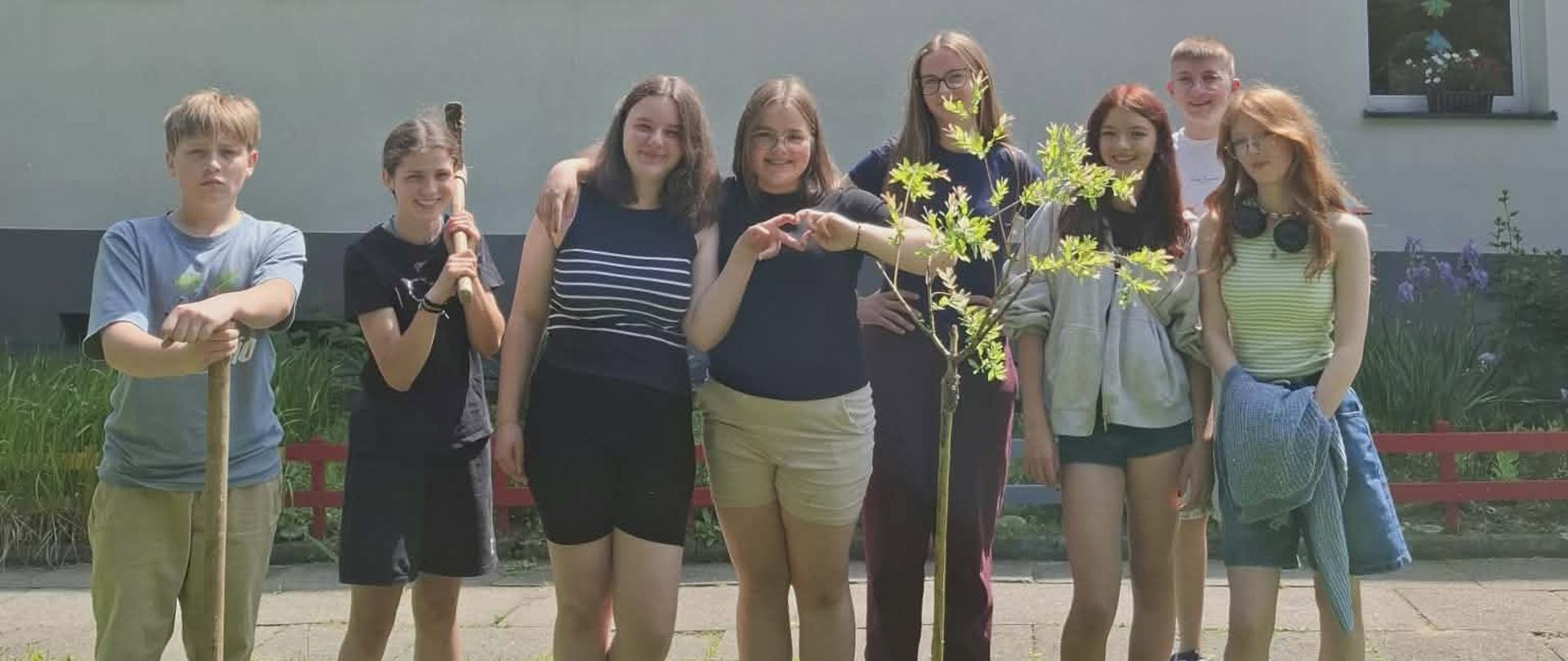 Klasa ósma stoi przed szkołą obok zasadzonego drzewka