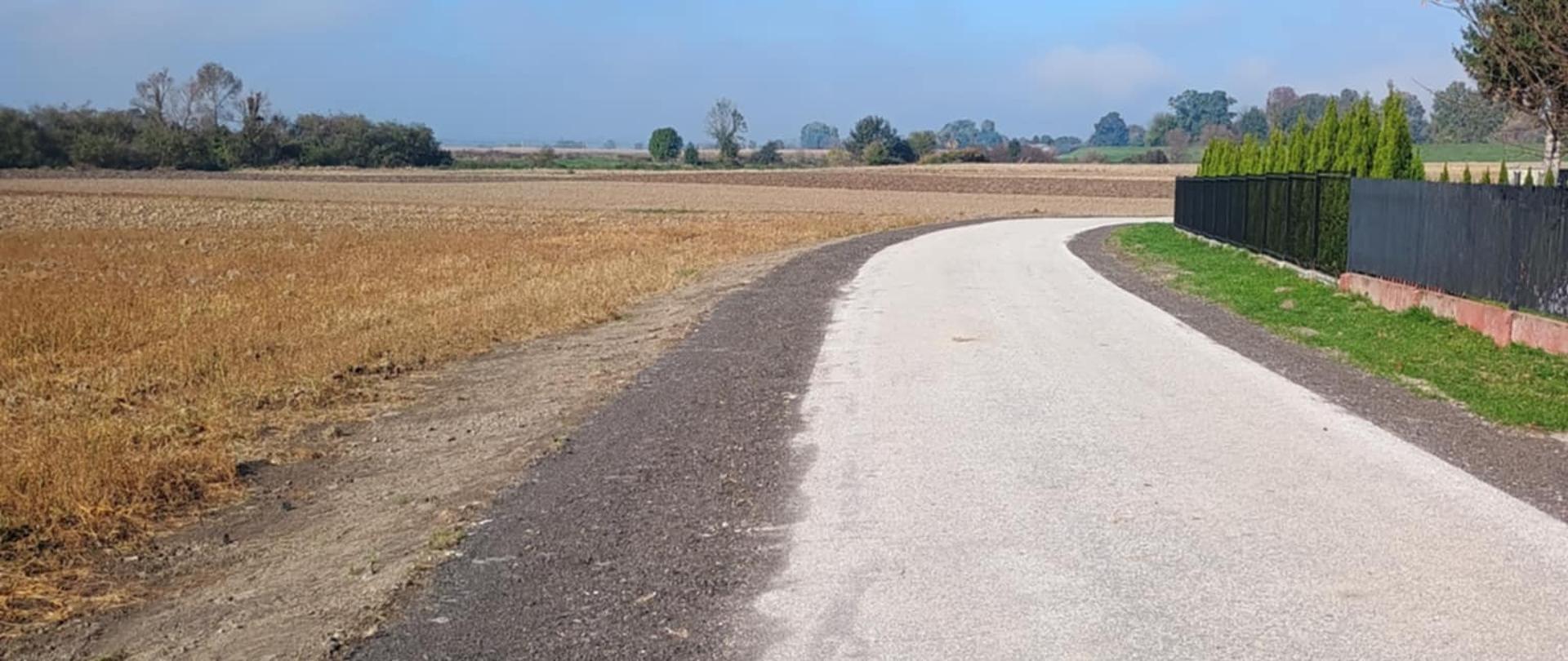 Zdjęcie przedstawia fragment drogi asfaltowej oraz pobocza z utwardzonego kamienia. Z lewej strony widać pola oraz drzewa w oddali. Z prawej strony widać fragment ogrodzenia.