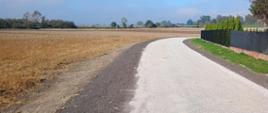 Zdjęcie przedstawia fragment drogi asfaltowej oraz pobocza z utwardzonego kamienia. Z lewej strony widać pola oraz drzewa w oddali. Z prawej strony widać fragment ogrodzenia.