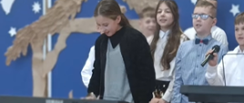 Dziewczynka w odświętnym stroju gra na keyboardzie, w tle inne dzieci śpiewają, jeden chłopiec trzyma w ręku mikrofon, stoi obok drugiego keyboardu