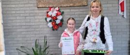 Dziewczynka w fioletowych spodniach i różowym bezrękawniku trzyma dyplom i nagrodę. Obok stoi Pani w czarnych spodniach, białej bluzce i czarnej kamizelce trzymając zająca - pracę plastyczną wykonaną przez uczennice.