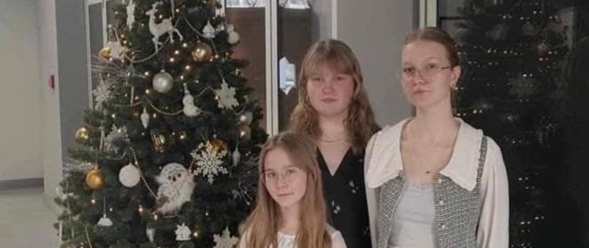 Trzy dziewczyny biorące udział w Międzynarodowym Festiwalu Kolęd i Pastorałek. W tle znajduje się przyozdobiona choinka.