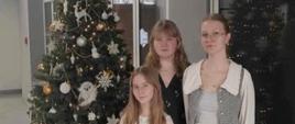 Trzy dziewczyny biorące udział w Międzynarodowym Festiwalu Kolęd i Pastorałek. W tle znajduje się przyozdobiona choinka.