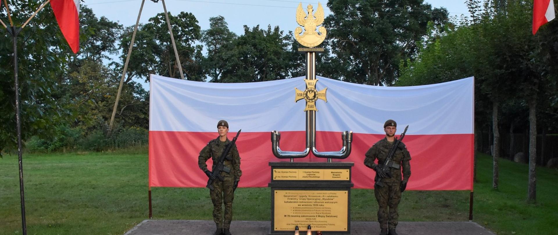 Dwóch żołnierzy stojących przy pomniku na warcie, za nimi rozwieszona flaga Polski.