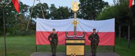 Dwóch żołnierzy stojących przy pomniku na warcie, za nimi rozwieszona flaga Polski.