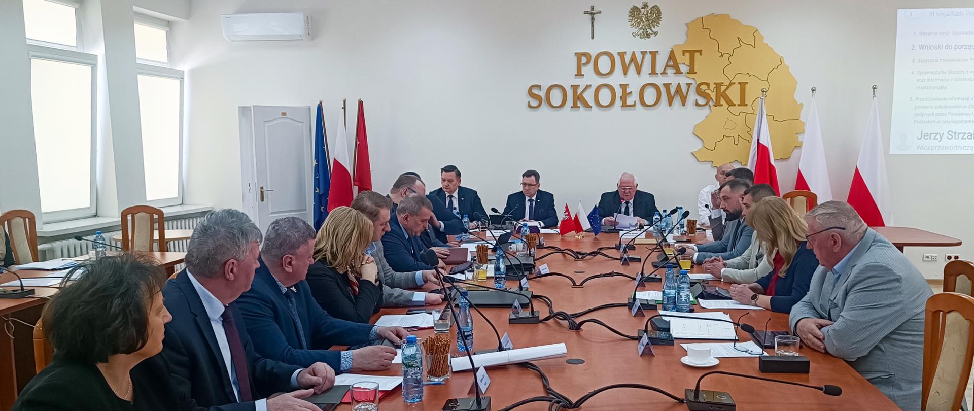 Na zdjęciu widok na salę oraz stół, przy którym toczą się obrady. Po jego obu stronach widzimy Radnych Rady Powiatu Sokołowskiego. Na przeciwległej ścianie widzimy duży przestrzenny napis Powiat Sokołowski wpisany w przestrzenny zarys granic powiatu. Nad całością krzyż katolicki oraz godło. W obu narożnikach widzimy flagi Polski, Unii Europejskiej oraz Mazowsza.