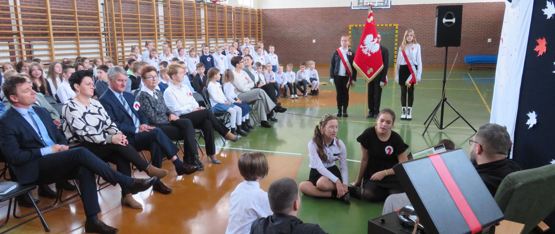 publiczność siedząca na krzesłach w sali gimnastycznej, troje dzieci z biało czerwonymi szarfami i czerwonym sztandarem z godłem Polski, grupa osób prawdopodobnie przedstawiająca inscenizację siedząca na podłodze, czarna walizka z czerwonym paskiem przez środek stojąca na stoliku