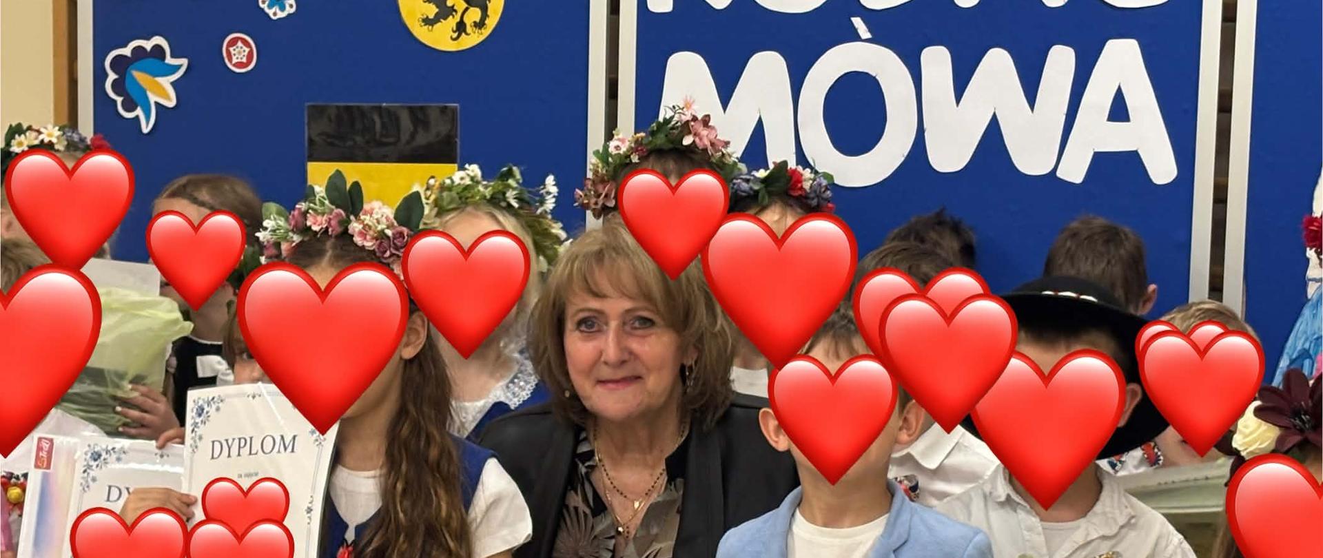 Na środku zdjęcia kobieta. Wokół niej stoją dzieci w kaszubskich strojach, twarze mają zasłonięte serduszkami. W rękach trzymają dyplomy i nagrody. W tle widać niebieską tablicę. 