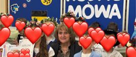 Na środku zdjęcia kobieta. Wokół niej stoją dzieci w kaszubskich strojach, twarze mają zasłonięte serduszkami. W rękach trzymają dyplomy i nagrody. W tle widać niebieską tablicę. 
